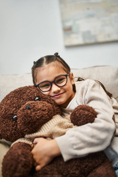 joyful girl in casual wear and eyeglasses hugging soft teddy bear and sitting on sofa in living room - Photo, Image