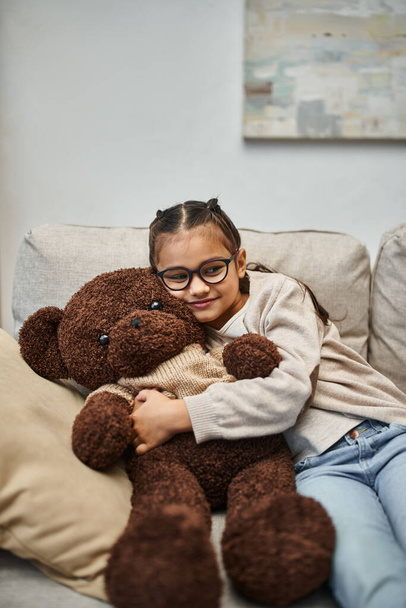 happy kid in casual wear and eyeglasses hugging soft teddy bear and sitting on sofa in living room - Photo, Image