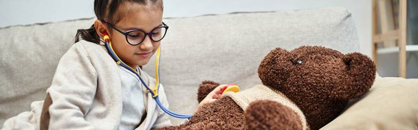 happy kid in casual wear and eyeglasses playing doctor with teddy bear on sofa in living room - Photo, Image