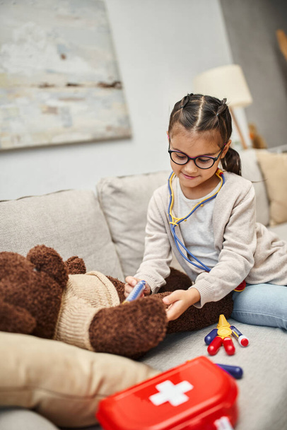 happy child in casual wear and eyeglasses playing doctor with teddy bear on sofa in living room - Photo, Image