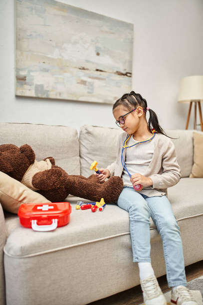 happy kid in casual wear and eyeglasses playing doctor with soft teddy bear on sofa in living room - Photo, Image