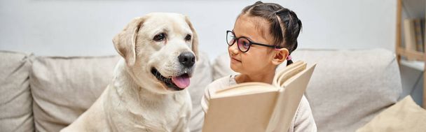 joyful girl in casual wear and eyeglasses reading book to labrador dog on in living room, banner - Photo, Image
