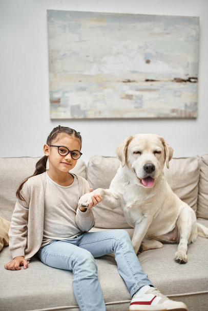 glückliches Mädchen im Grundalter mit Brille, die Pfote eines Labradors haltend und auf dem Sofa im Wohnzimmer sitzend - Foto, Bild