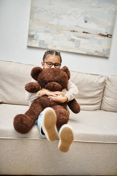 imádnivaló általános iskolás lány szemüvegben kezében játékmackó és ül a kanapén a nappaliban - Fotó, kép