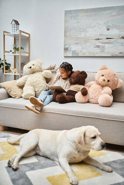 rozkošná dívka sedí na pohovce s měkkými medvídky a čtení knihy v blízkosti labrador v obývacím pokoji - Fotografie, Obrázek