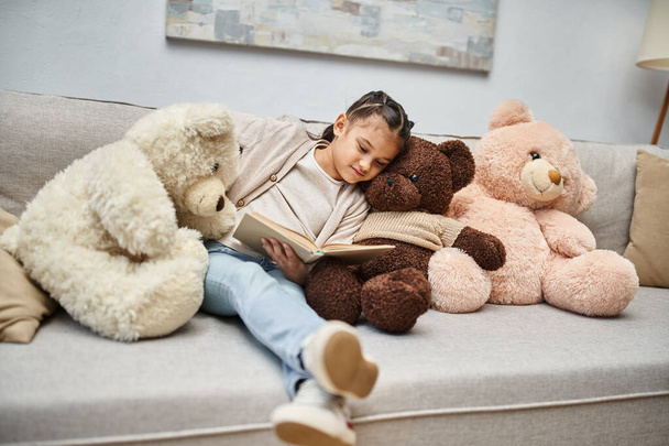 adorable chica sentada en el sofá con suaves osos de peluche y libro de lectura en la sala de estar moderna - Foto, Imagen