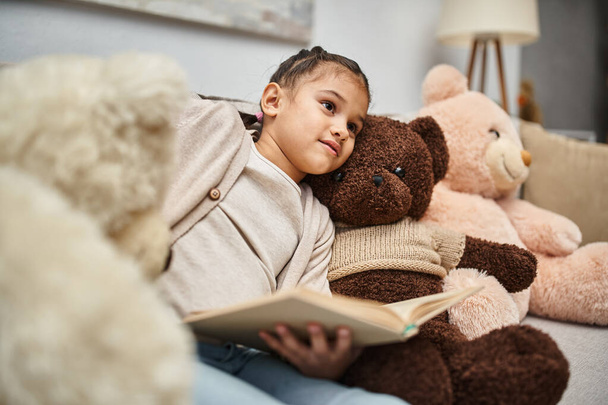 cute girl sitting on comfortable sofa with soft teddy bears and reading book in modern living room - Photo, Image