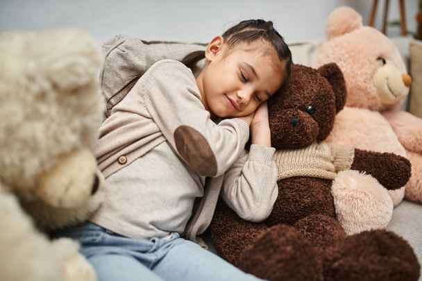 adorável menina idade elementar dormindo entre ursos de pelúcia macios no sofá na sala de estar moderna - Foto, Imagem