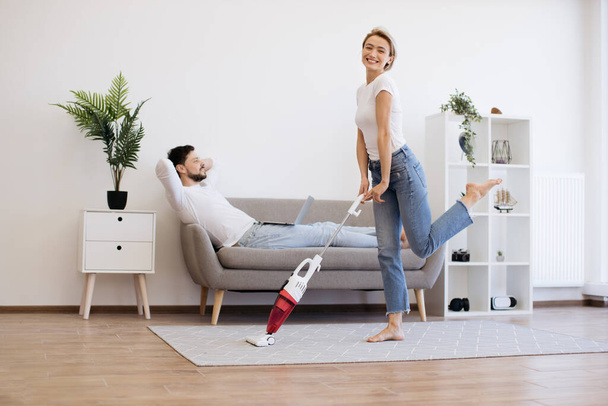 Cheerful caucasian woman in casual wear dancing with cordless vacuum cleaner during chores while focused husband sitting on couch and working on laptop. Lifestyles of modern young family. - 写真・画像