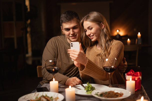 Περιήγηση στο διαδίκτυο κατά τη διάρκεια ρομαντικό δείπνο ημερομηνία στο σπίτι. Ευτυχισμένο ζευγάρι ψάχνει φωτογραφίες στο smartphone κατά τη διάρκεια του εορτασμού Ημέρα του Αγίου Βαλεντίνου. Αντιγραφή χώρου - Φωτογραφία, εικόνα