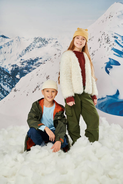 vertical shot of little boy sitting on snow near cute standing girl, stylish outfit, winter fashion - Photo, Image