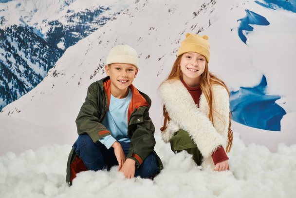 happy preadolescent boy and girl in warm attire sitting on snow and looking at camera, fashion - Photo, Image