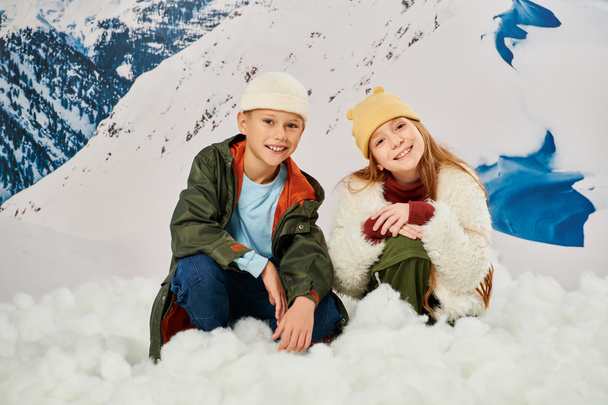 fröhliche kleine Jungen und Mädchen sitzen auf Schnee in warmen, stylischen Outfits und lächeln in die Kamera, Mode - Foto, Bild