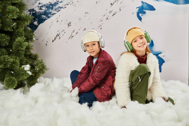 cute boy and girl in stylish winter outfits with headsets sitting on snow with mountain backdrop - Photo, Image