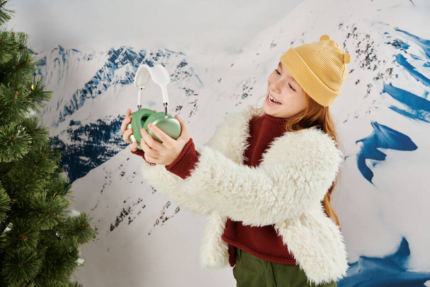 szczęśliwy nastolatek dziewczyna w zimie stylowy strój obok jodły patrząc na jej słuchawki, koncepcja mody - Zdjęcie, obraz