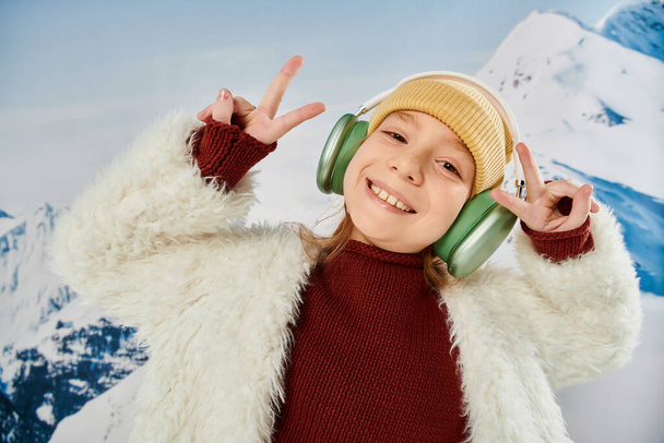 portrait of preteen girl with headset smiling joyfully at camera showing peace sign, fashion concept - Photo, Image
