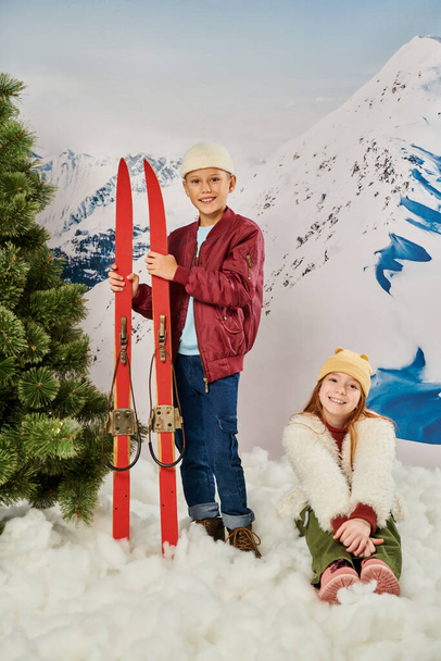 vertical shot of little boy standing and holding skis next to cute girl sitting on snow, fashion - Photo, Image