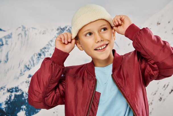 cheerful little boy in stylish attire putting on his trendy beanie hat looking away, fashion concept - Photo, Image