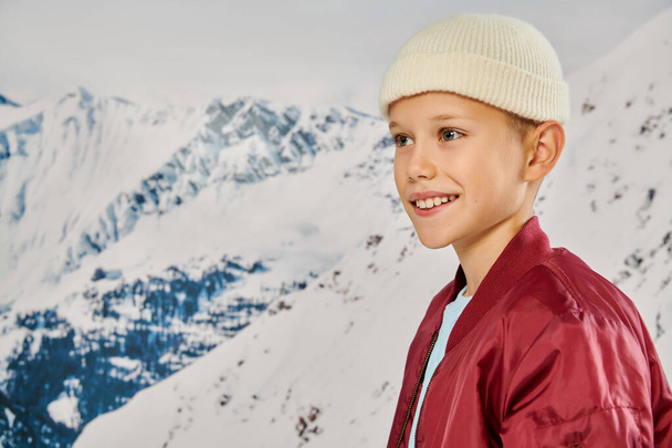 portrait of jolly preteen boy in beanie hat looking away with snowy mountain on backdrop, fashion - Photo, Image