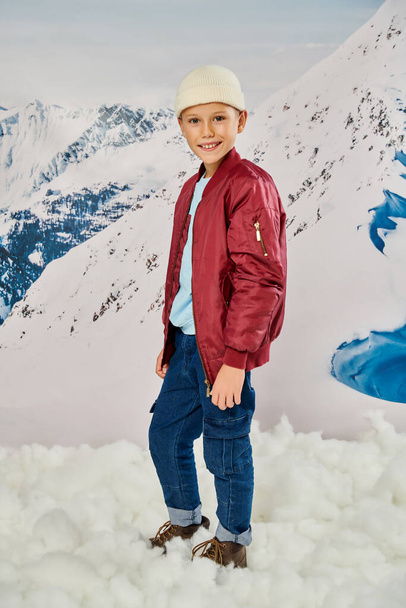vertical shot of little boy in stylish warm clothes standing on snow and smiling at camera, fashion - Photo, Image