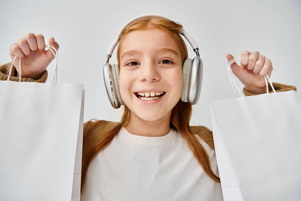radosna dziewczynka z nowoczesnym zestawem słuchawkowym pozowanie z prezentowanymi torbami na szarym tle, koncepcja mody - Zdjęcie, obraz