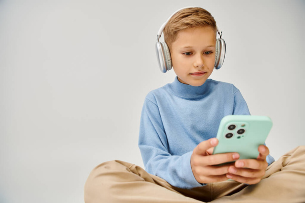 милый мальчик-подросток в синей модной толстовке сидит на полу с наушниками и смотрит в телефон - Фото, изображение
