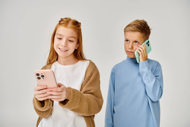 сосредоточенный маленький мальчик разговаривает по телефону рядом со своим веселым другом глядя на ее мобильный, мода концепции - Фото, изображение