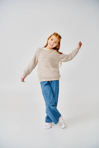 веселая девочка-подросток в трикотажном свитере и джинсах позирует в движении на сером фоне, мода - Фото, изображение