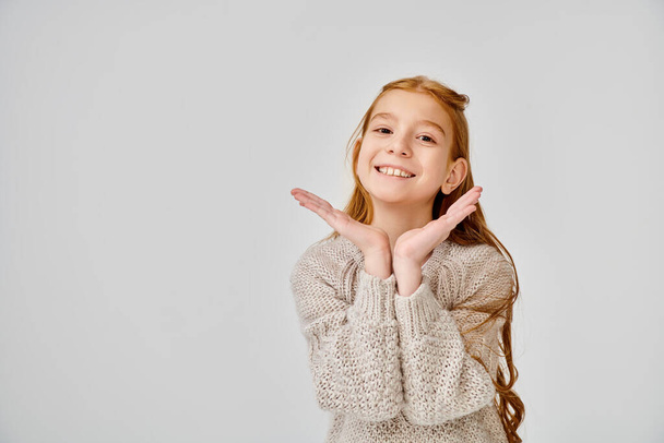 красивая маленькая девочка с рыжими волосами в обычном вязаном свитере, смотрящая на камеру с руками под лицом - Фото, изображение
