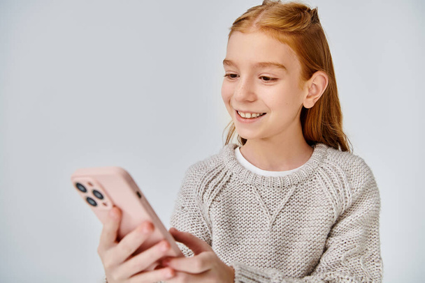 веселая девочка-подросток с рыжими волосами, радостно смотрящая на свой телефон на сером фоне, концепция моды - Фото, изображение
