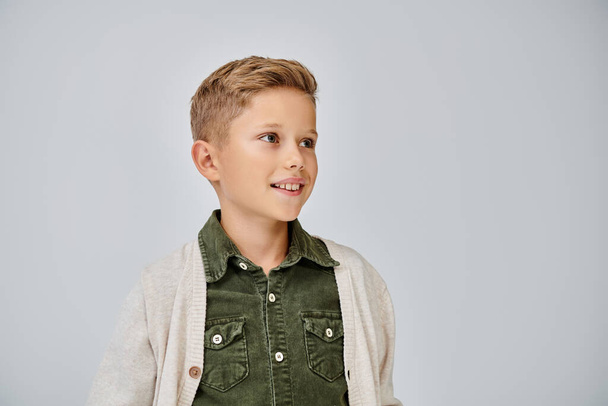 vrolijk jongetje in warme casual vest glimlachend en wegkijkend op grijze achtergrond, fashion concept - Foto, afbeelding