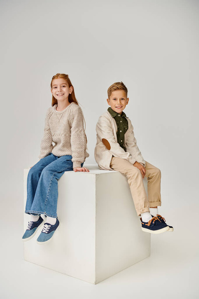 vertical shot of two little children in casual trendy attires sitting on cube and smiling at camera - Photo, Image