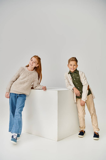 vertikale Aufnahme von kleinen stilvollen Freunden, die sich auf einen weißen riesigen Würfel vor grauem Hintergrund lehnen, Modekonzept - Foto, Bild