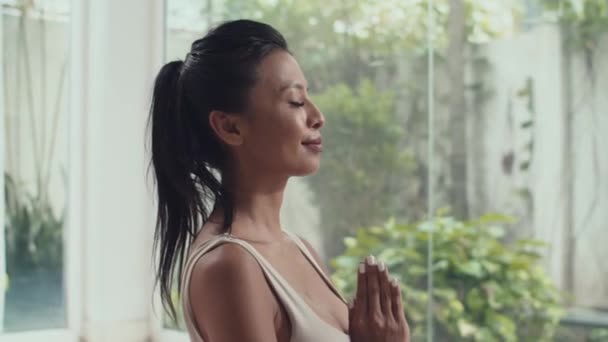 Tilt down shot of Asian woman meditating with eyes closed while doing yoga at home - Footage, Video