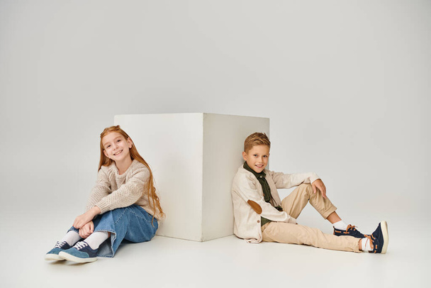 χαρούμενα προεφηβικά παιδιά που κάθονται στο πάτωμα δίπλα σε λευκό κύβο και χαμογελούν στην κάμερα, μόδα - Φωτογραφία, εικόνα
