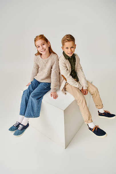cheerful preteen boy and girl in warm trendy clothes sitting on cube and smiling at camera, fashion - Photo, Image