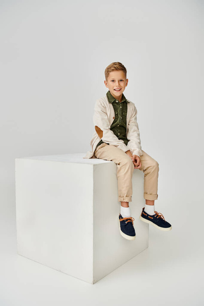 вертикальный снимок радостного мальчика в модном кардигане, сидящего на белом кубе и улыбающегося в камеру - Фото, изображение