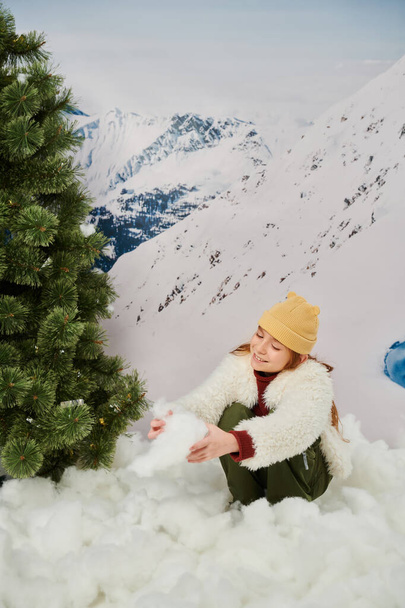 cheerful red haired little girl playing with snow next to fir tree with mountain on backdrop - Photo, Image