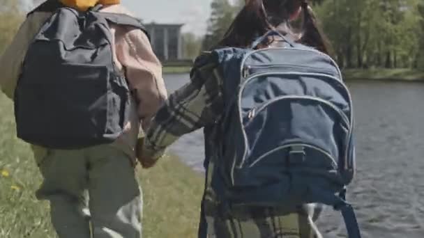 Pohled zezadu na dvě holčičky s batohy, které se drží za ruce a jdou po břehu řeky v parku po škole - Záběry, video