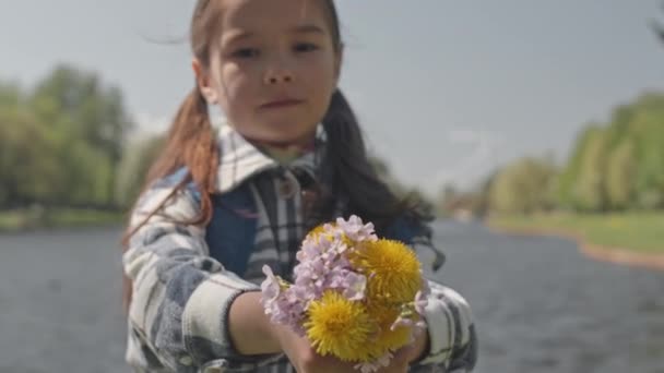 Zoom out portrait shot of little girl holding bouquet of wildflowers in hands outstretched to camera and posing by river in park - Footage, Video
