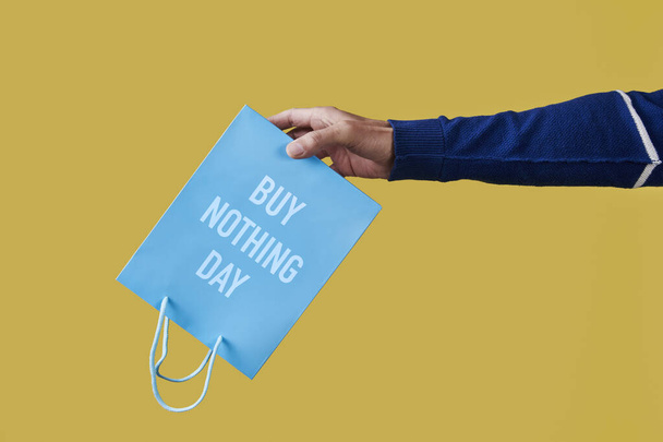 primer plano de un hombre sosteniendo una bolsa de papel azul, con el texto comprar nada día escrito en él, al revés sobre un fondo amarillo - Foto, Imagen