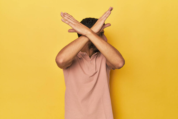 Νεαρός Ισπανόφωνος με κίτρινο φόντο κρατώντας δύο χέρια σταυρωμένα, έννοια της άρνησης. - Φωτογραφία, εικόνα