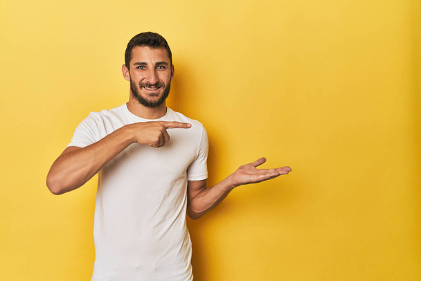 Νεαρός Ισπανόφωνος σε κίτρινο φόντο ενθουσιασμένος κρατώντας ένα χώρο αντίγραφο στην παλάμη. - Φωτογραφία, εικόνα