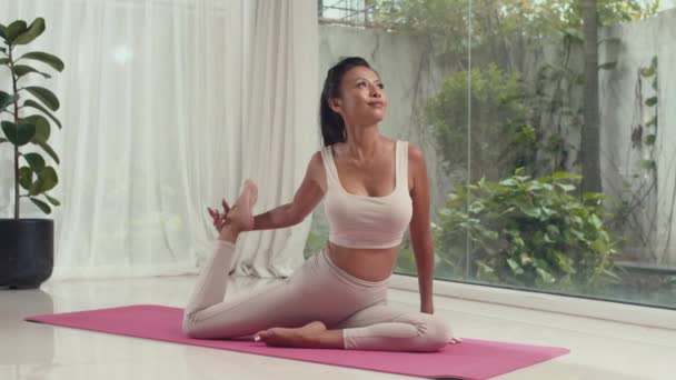 Longo tiro de jovem mulher flexível esticando pernas, braços e corpo ao fazer ioga no tapete em casa, asana - Filmagem, Vídeo