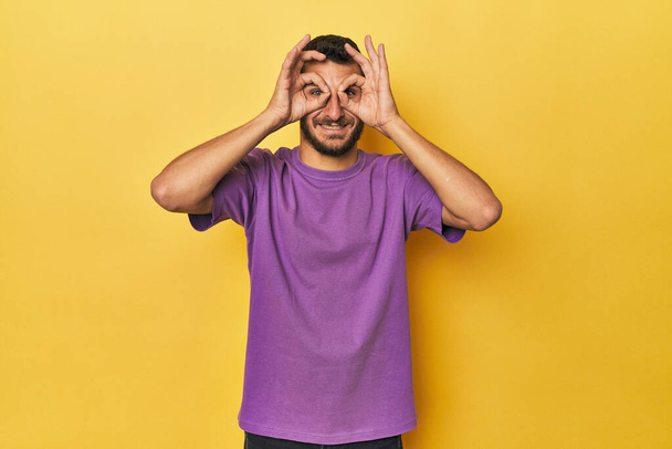 Молодой латиноамериканец на жёлтом фоне показывает знак "хорошо" над глазами - Фото, изображение