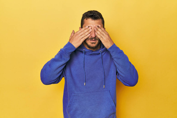 Jeune homme hispanique sur fond jaune peur couvrant les yeux avec les mains. - Photo, image