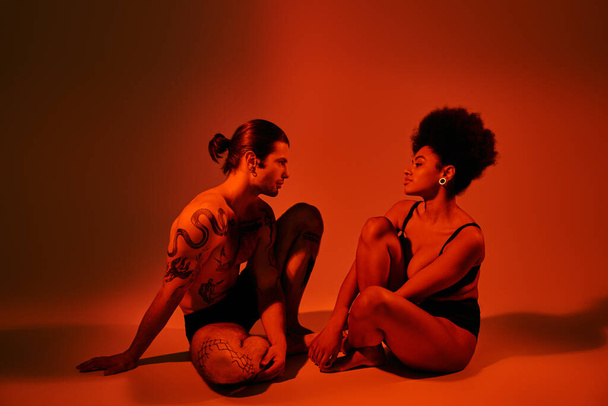 aantrekkelijk Afrikaans amerikaans vrouw kijken naar haar getatoeëerde vriend terwijl beide zitten op de vloer - Foto, afbeelding