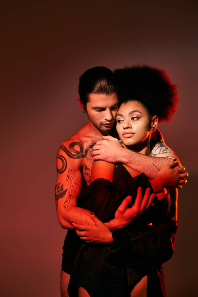 εμφανίσιμος άντρας με τατουάζ που αγκαλιάζει με αγάπη την Αφροαμερικανή φιλενάδα του από πίσω, σέξι ζευγάρι - Φωτογραφία, εικόνα