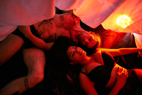 seductora pareja multicultural acostada sensualmente bajo sábanas rodeada de luces - Foto, Imagen