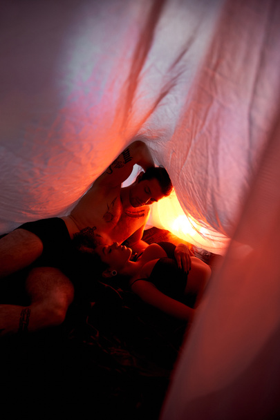 σέξι πολυπολιτισμικό ζευγάρι με εσώρουχα ξαπλωμένα αισθησιακά μαζί κάτω από σεντόνι που περιβάλλεται από φώτα - Φωτογραφία, εικόνα